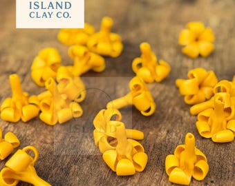 Pode incluir: Uma coleção de pequenos componentes de argila amarela brilhante em forma de flor espalhados sobre uma superfície de madeira. Os componentes têm um design detalhado e tridimensional. O logotipo da Island Clay Co. está no canto superior esquerdo.