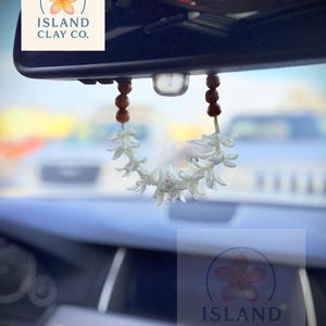 Peut inclure: Un d&eacute;sodorisant de voiture en forme de fleur blanche suspendu au r&eacute;troviseur d'une voiture. Le d&eacute;sodorisant est compos&eacute; de fleurs blanches et de perles en bois marron. Le logo Island Clay Co. avec une fleur est &eacute;galement visible.