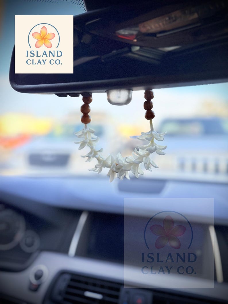 Peut inclure: Un d&eacute;sodorisant de voiture en forme de fleur blanche suspendu au r&eacute;troviseur d'une voiture. Le d&eacute;sodorisant est compos&eacute; de fleurs blanches et de perles en bois marron. Le logo Island Clay Co. avec une fleur est &eacute;galement visible.