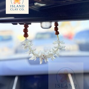 Peut inclure: Un d&eacute;sodorisant de voiture en forme de fleur blanche suspendu au r&eacute;troviseur d'une voiture. Le d&eacute;sodorisant est compos&eacute; de fleurs blanches et de perles marron. Le logo Island Clay Co. est visible.