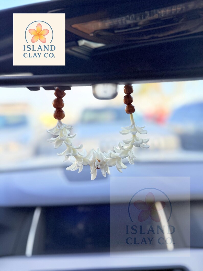 Peut inclure: Un d&eacute;sodorisant de voiture en forme de fleur blanche suspendu au r&eacute;troviseur d'une voiture. Le d&eacute;sodorisant est compos&eacute; de fleurs blanches et de perles marron. Le logo Island Clay Co. est visible.