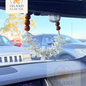 Peut inclure: Un d&eacute;sodorisant de voiture avec une guirlande de fleurs blanches et des perles en bois, suspendu au r&eacute;troviseur. Le tableau de bord et le logo "ISLAND CLAY CO." sont visibles. L'heure est 15h26.