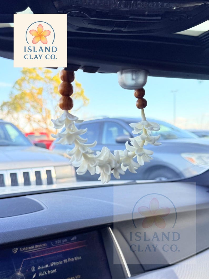 Peut inclure: Un d&eacute;sodorisant de voiture avec une guirlande de fleurs blanches et des perles en bois, suspendu au r&eacute;troviseur. Le tableau de bord et le logo "ISLAND CLAY CO." sont visibles. L'heure est 15h26.