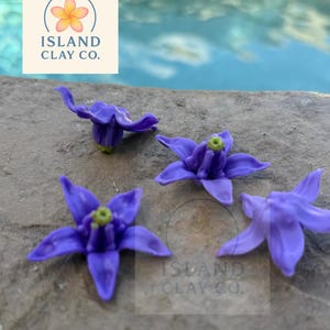 May include: Four small, purple flower-shaped clay pieces with green centers are arranged on a gray surface. The flowers have five petals each. The Island Clay Co. logo is visible in the background.