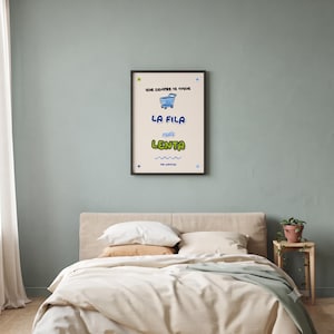 May include: A framed wall art print with a cream background featuring a blue shopping cart illustration and the Spanish text: Que siempre te toque LA FILA más LENTA. Below the text is a blue wavy line and the words THE UNIVERSE with small stars.