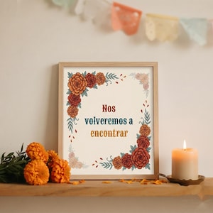 May include: A framed print featuring the text "Nos volveremos a encontrar" in the center, surrounded by a decorative border of orange and red marigold flowers and green leaves on a cream background, presented in a light wood frame.