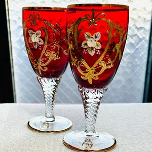 Puede incluir: Dos copas de cristal rojo rubí con motivos de corazón en filigrana dorada y detalles florales blancos pintados a mano. Cada copa tiene un tallo transparente retorcido y una base circular con un borde dorado. Un logo dorado es visible.