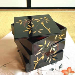 Puede incluir: Una pila de cuatro cajas lacadas en negro con diseños botánicos en pan de oro. Las cajas son cuadradas y tienen un acabado brillante. La tapa de la caja superior está ligeramente entreabierta, revelando un interior rojo. Las cajas están apiladas sobre una tela estampada floral.