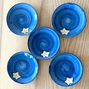 May include: Five small, round, blue ceramic plates with a spiral design. Each plate has a white star-shaped accent. The plates are arranged on a light-colored wooden surface.