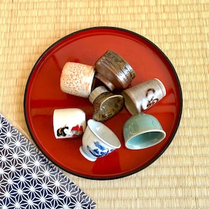 May include: A selection of small ceramic sake cups in various colours and patterns, arranged on a bright red lacquered tray. The cups include designs with floral, abstract, and traditional Japanese motifs. The tray sits on a woven mat.