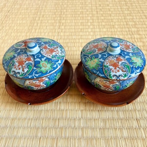 May include: Two matching blue and white ceramic bowls with lids, decorated with orange, green, and white floral designs. Each bowl rests on a small, round, dark wooden saucer. The background is a woven straw mat.