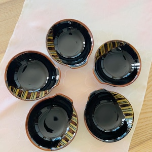 May include: A set of five small ceramic bowls with a glossy black interior and a textured, earthy brown rim featuring a decorative pattern of vertical yellow and dark brown stripes. Each bowl has a distinct, slightly irregular circular shape.