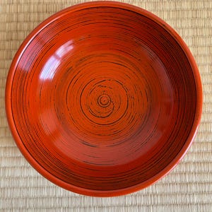 以下が含まれることがあります： 光沢のある仕上がりの丸い赤い漆器のボウル。ボウルは、内側に渦巻く同心円状の黒い線を特徴としています。ボウルは、織られた薄茶色のマットの上に置かれています。