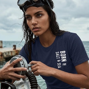 May include: A navy blue short-sleeved t-shirt featuring the white text BORN OF SALT & SEA printed vertically on the chest. The shirt is worn with a black diving mask positioned on the forehead and is paired with a metallic scuba diving tank.