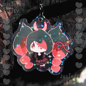 May include: A clear, iridescent acrylic charm featuring a cartoon character with black hair, red accents, and a red bow. The charm is attached to a metal chain. The text "Juliette Lily" is visible.