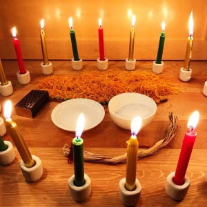 May include: A collection of lit candles in red, green, and gold, set in white ceramic holders. The candles are arranged around a wooden surface with a scattering of orange material, two white bowls, and a small wooden box. The scene is illuminated by the candle flames.