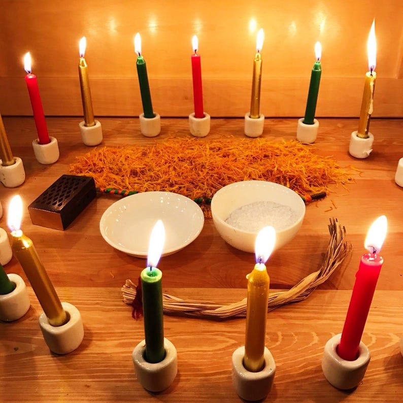 May include: A collection of lit candles in red, green, and gold, set in white ceramic holders. The candles are arranged around a wooden surface with a scattering of orange material, two white bowls, and a small wooden box. The scene is illuminated by the candle flames.