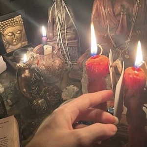 May include: A dimly lit scene featuring several Buddha statues and lit candles. The candles are orange and red, with bright flames. A hand is reaching towards the candles. Other objects include a framed Buddha image and a small glass jar.