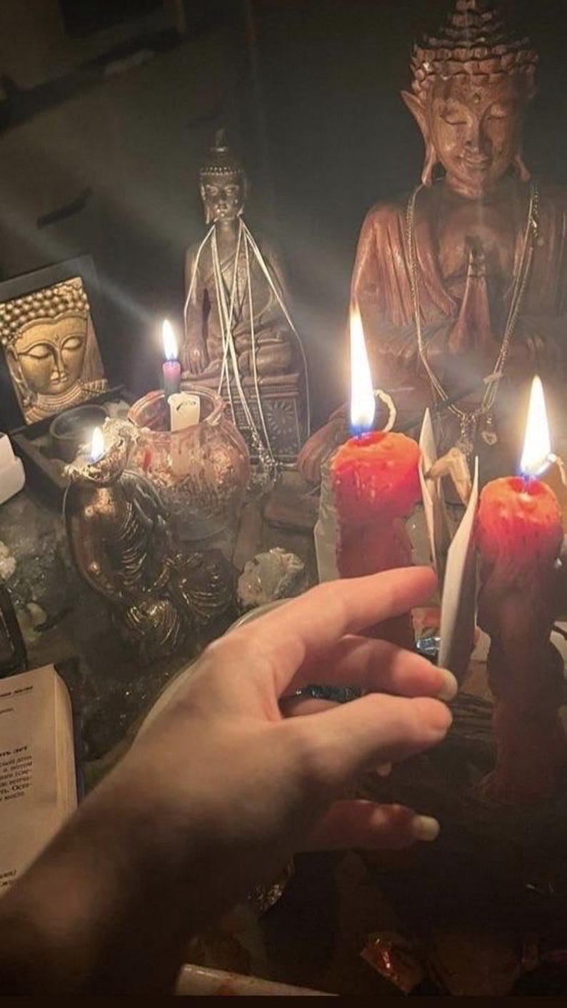 May include: A dimly lit scene featuring several Buddha statues and lit candles. The candles are orange and red, with bright flames. A hand is reaching towards the candles. Other objects include a framed Buddha image and a small glass jar.