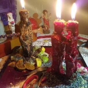May include: Lit candles with bright flames. One candle is brown and textured, the other two are red. The candles are surrounded by herbs, fruit slices, and small figurines. A pair of scissors and a knife are also visible.