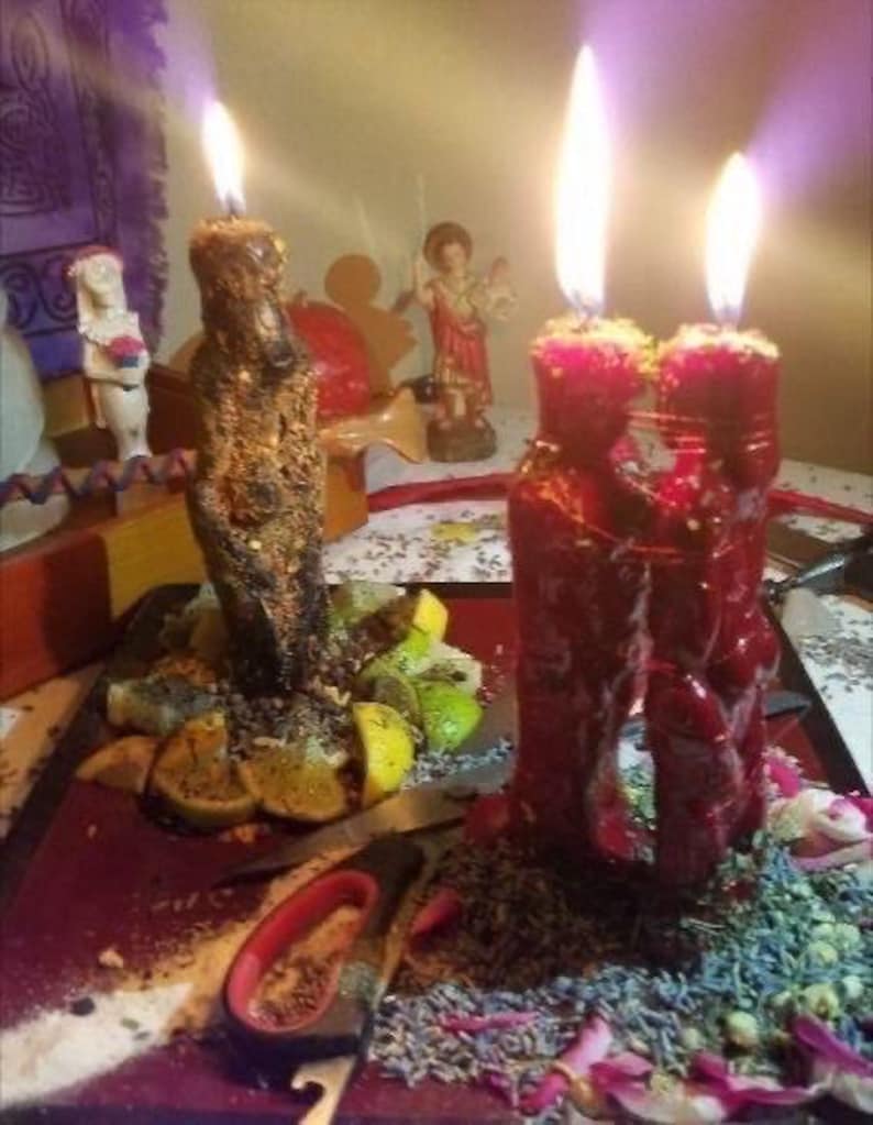 May include: Lit candles with bright flames. One candle is brown and textured, the other two are red. The candles are surrounded by herbs, fruit slices, and small figurines. A pair of scissors and a knife are also visible.