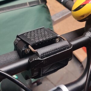 Könnte beinhalten: Schwarzer Fahrradlenker mit einem schwarzen, lederartigen Zubehör, das mit einem Riemen und Metallbeschlägen befestigt ist. Eine gelbe Fahrradklingel mit roten Akzenten ist sichtbar. Eine grüne Tasche ist im Hintergrund.