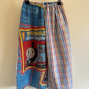 May include: Trousers featuring a split design with one leg displaying a Thomas the Tank Engine graphic print on a blue background with red accents and the text Thomas Blue Engine, and the other leg featuring a blue, red, and yellow plaid pattern.