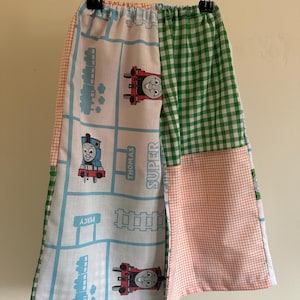 May include: Trousers featuring a patchwork design with a white fabric panel printed with blue train tracks and Thomas the Tank Engine characters labelled Thomas and Percy, alongside green and white gingham and orange and white checkered fabric panels.