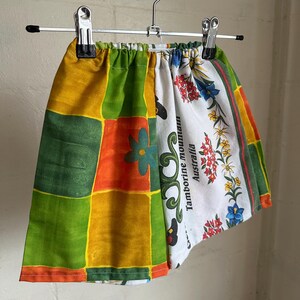 May include: A pair of shorts made from repurposed fabric featuring a patchwork of green, yellow, and orange squares on one side and a white panel with floral patterns and the text Tamborine Mountain Australia on the other side.