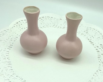 May include: Two small, pink ceramic vases with a bulbous base and a narrow neck. The interior of the vases is white. The vases are displayed on a white, lacy doily.