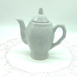 May include: A small, light gray ceramic teapot with a lid and handle. The teapot has a crackle glaze finish, giving it a textured appearance. The spout is curved, and the handle is a loop shape. The teapot is sitting on a white surface.