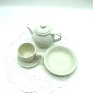 May include: A small, white ceramic tea set, including a teapot, teacup with saucer, and a small plate. The set is arranged on a white, lace-edged doily against a white background. The teapot has a lid and a curved handle.