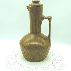 May include: A brown ceramic pitcher with a lid and handle. The pitcher has vertical ribbed detailing and a rounded base. It is sitting on a white paper doily.