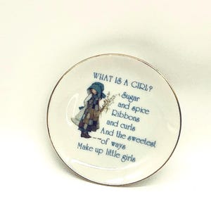 May include: A small, round, white decorative plate with a gold rim. The plate features a blue illustration of a girl and text that reads "WHAT IS A GIRL? Sugar and spice Ribbons and curls And the sweetest --of ways Make up little girls."