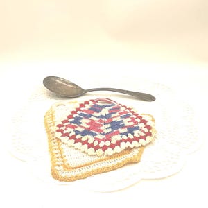 May include: A stack of two crocheted pot holders with a spoon on a white doily. The top pot holder features a geometric pattern in red, white, and blue. The bottom pot holder has a yellow border. The spoon is silver.