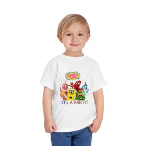 May include: A white toddler t-shirt featuring the text "Yo Gabba Gabba!" above a group of colorful cartoon characters. Below the characters, the shirt reads "It's a Party!" The shirt is worn by a young child.