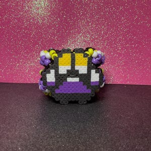 May include: A pixelated bracelet in a non-binary pride flag design. The bracelet features a black base with yellow, white, purple, and gray accents. The design is made of small, fused beads, creating a textured appearance. The bracelet is a unique accessory.