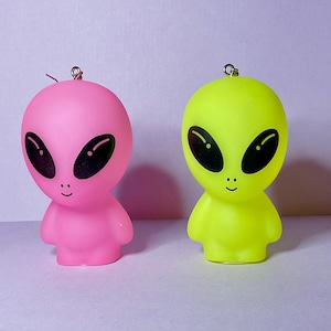 Op de afbeelding: Twee buitenaardse figuren. Eén is felroze, de andere neon groen. Beide hebben grote zwarte ovale ogen en kleine glimlachjes. Elk heeft een kleine metalen lus aan de bovenkant. De achtergrond is lichtpaars.