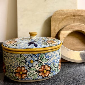 May include: A round ceramic container with a lid, featuring a hand-painted floral pattern in blue, orange, and green on a white background. Beside it are a light-colored marble cutting board with a leather strap and a wooden board with a metal blade.