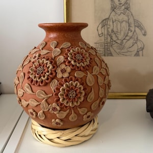 May include: A round terracotta vase with a narrow neck, featuring raised relief floral and leaf patterns in shades of brown and tan. The vase sits on a circular, woven light-colored reed base, showcasing intricate clay craftsmanship.