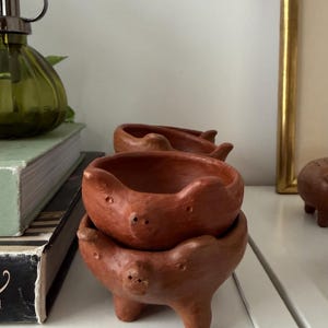 May include: A stack of two small, reddish-brown terracotta bowls shaped like pigs, each featuring molded ears, a snout with two nostrils, and small eyes. The bowls are rounded with three short, sturdy legs, showcasing a natural clay texture.