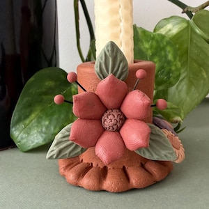 May include: A cylindrical terracotta candle holder featuring a sculpted, matte red flower with a textured center, surrounded by sage green leaves and small red spheres on thin wire stems, with a tall, cream-colored, textured taper candle inserted.