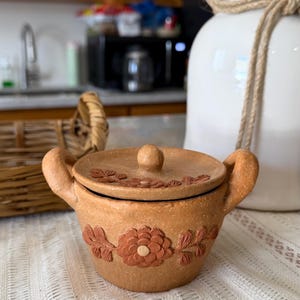 Könnte beinhalten: Ein kleiner, runder Tontopf in Terrakotta-Farbe mit passendem Deckel und zwei seitlichen Griffen. Die Außenseite ist mit einem erhabenen, strukturierten Blumenmuster verziert, das eine zentrale Blüte mit Blättern zeigt, auch der Deckel hat ein Motiv.