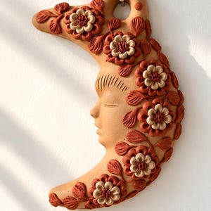 Pode incluir: Uma decoração de parede em forma de meia-lua feita de argila cor de terracota. A peça apresenta um rosto humano esculpido de perfil com olhos fechados, adornado com motivos florais e de folhas em relevo em tons de terracota e creme.