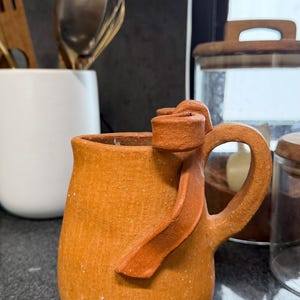 May include: A terracotta-colored ceramic pitcher with a textured, matte finish. The pitcher features a rounded body and a curved handle with a decorative, ribbon-like clay accent attached to the top rim, extending downward along the side.