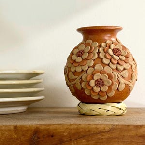 Puede incluir: Un jarrón redondo de terracota con flores de arcilla tridimensionales que presentan pétalos superpuestos y centros texturizados. El jarrón descansa sobre una base circular trenzada de color claro, con un acabado natural y terroso.