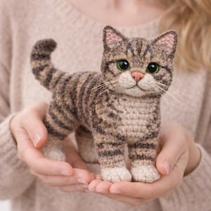 Op de afbeelding: Een handgemaakte gehaakte kattenfiguur in bruin- en beigetinten, met groene ogen en een roze neus. De kat heeft gestreepte markeringen en wordt in open handen gehouden. Een charmant, grillig kattenbeeld.