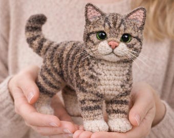 May include: A handmade crochet cat figurine in shades of brown and tan, with green eyes and a pink nose. The cat has striped markings and is held in open hands. A charming, whimsical cat sculpture.