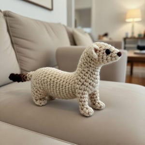Könnte beinhalten: Gehäkelte Frettchenfigur aus cremefarbenem Garn. Das Frettchen hat schwarze Augen, eine braune Nase und einen Schwanz mit braunen und cremefarbenen Streifen. Das handgefertigte Tier steht auf einem beigen Sofa.