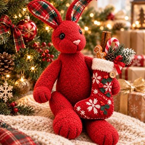Könnte beinhalten: Ein roter Plüschhase mit langen Ohren, die ein grün-rotes Karomuster aufweisen. Der Hase hält einen roten Weihnachtsstrumpf, der mit weißen Schneeflocken, grünen Stechpalmenblättern, einem Lebkuchenmann, einer Zuckerstange und einer Zimtstange verziert ist.